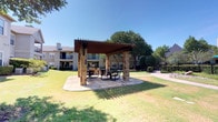 The grill and picnic area is a well-appointed outdoor amenity space designed for gathering and outdoor dining. At its centerpiece stands a substantial pergola constructed with a decorative wood-lattice roof and supported by sturdy natural stone columns, providing partial shade while maintaining an open, airy atmosphere. The pergola shelters multiple round tables paired with metal mesh chairs, offering comfortable seating for residents to dine and socialize outdoors.

The patio surface beneath the pergola features a stone tile design, adding a refined, textured look to the space. Adjacent to the seating area, a stone-clad grill station provides a dedicated space for outdoor cooking. The pergola's ironwork wall brackets add a decorative architectural detail that complements the natural stone elements throughout.

Surrounding the covered area, expansive, well-maintained green lawns extend outward, dotted with mature shade trees that contribute to the space's inviting outdoor ambiance. Concrete walkways connect the area to the broader grounds, which are bordered by ornamental iron fencing and decorative rock landscaping features. Views of the surrounding buildings and additional umbrella-shaded seating areas are visible from the space, reinforcing its role as a central outdoor hub within the community.