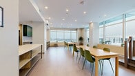 This contemporary 760 square foot workspace, located at 300 Place Charles-le Moyne in Longueuil, Quebec, offers a versatile and efficient layout. The space is characterized by its open floor plan, which promotes collaboration and flexibility. Large windows line the exterior walls, flooding the area with natural light and providing panoramic city views.

The interior features a neutral color palette with pops of teal and green, creating a fresh and inviting atmosphere. A well-equipped kitchenette area includes sleek cabinetry, a sink, and necessary appliances. Adjacent to this is a multi-functional island with built-in storage, ideal for casual meetings or additional workspace.

The main area accommodates multiple wooden tables with colorful chairs, suitable for various work arrangements or group discussions. A separate enclosed meeting room with glass walls maintains visual connectivity while offering privacy when needed.

Recessed lighting throughout ensures even illumination, complementing the abundant natural light. The space is climate-controlled for comfort, with visible air conditioning units. Safety features such as fire extinguishers and emergency exit signs are strategically placed.

Overall, this space combines functionality with modern design, creating an environment conducive to productivity and teamwork in a professional setting.