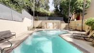 This outdoor amenity space is designed for relaxation and recreation, centered around a generously sized swimming pool with clear, turquoise water. The pool features a distinctive shape with a dedicated wading area, tile-trimmed edges, and convenient pool entry steps with handrails. Surrounding the pool is a wide, open concrete deck furnished with lounge chairs and chaise lounges, offering ample space for sunbathing and leisure.

A dedicated poolside lounge area is equipped with a shaded dining table and chairs beneath a large patio umbrella, ideal for outdoor meals or casual gatherings. Adjacent to this seating area is a built-in BBQ grill station set against a stone-accented wall, providing a convenient outdoor cooking setup surrounded by lush, mature landscaping.

Changing room or restroom facilities are accessible from the pool deck, housed in a light-colored utility structure at the far end of the pool area. The entire space is enclosed by a chain-link security fence, with tall, mature trees providing natural shade and a sense of seclusion. Verdant plantings line the perimeter, including ornamental shrubs, tropical foliage, and climbing vines along ironwork fencing, creating a private, garden-like atmosphere throughout the outdoor space.