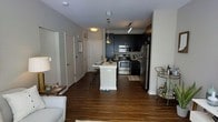 This single-floor residence showcases a thoughtfully designed open-concept layout that seamlessly connects the living and kitchen areas. Rich, dark hardwood flooring extends throughout the space, providing a warm contrast to the light-toned walls and white kitchen island.

The kitchen is a standout feature, equipped with stainless steel appliances including a range, microwave, and refrigerator. Striking dark navy cabinetry lines the walls, offering bold contrast against the white peninsula, which doubles as a breakfast bar with seating for two. Pendant lighting with industrial-style fixtures hangs above the island, complementing the track lighting overhead.

The adjacent living area is visible from the kitchen, reinforcing the open flow of the space. A bar cart near the living area adds a stylish, functional touch. Doors along the entry corridor suggest access to additional rooms, maintaining a clean and organized layout. The overall ambiance is modern and contemporary, blending dark and light tones with practical fixtures to create an inviting yet sophisticated atmosphere.