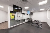 This fitness center is designed to support a comprehensive range of workout routines within a clean, modern environment. The space is finished with a neutral white and grey palette, dark rubberized flooring, and a suspended tile ceiling with recessed panel lighting that ensures consistent, even illumination throughout.

The cardio zone houses multiple Matrix-brand treadmills and elliptical trainers, as well as stationary bikes, providing ample options for aerobic training. Adjacent to this area, a dedicated free weights section features a full rack of dumbbells arranged in ascending order, paired with adjustable flat benches for a variety of strength exercises. Large floor-to-ceiling mirrors line the walls in this zone, enhancing the sense of space and allowing users to monitor their form.

A functional training area accommodates a multi-station cable resistance machine alongside stability balls stored on a vertical rack, supporting both strength and flexibility training. Several wall-mounted flat-screen televisions are positioned throughout the space, keeping users entertained during workouts. A bold, colorful mural depicting athletic silhouettes serves as an energizing focal point, adding a dynamic visual element to the otherwise minimalist interior. The overall layout is open and well-organized, allowing for comfortable circulation between equipment zones.