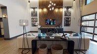 This single-floor amenity space showcases a striking industrial-modern design defined by soaring double-height ceilings and an open, flowing layout. The main gathering area centers around a full-size billiards table set atop a herringbone-patterned area rug, surrounded by wood-look tile flooring that extends throughout the space. A dramatic lounge area features a textured bamboo accent wall backlit by warm recessed lighting, serving as the backdrop for a wall-mounted television flanked by black-and-white urban photography. The seating arrangement includes a generous sectional sofa, an industrial-style console table with riveted metal detailing, and wire-frame floor lamps that reinforce the space's urban aesthetic.

Floor-to-ceiling glass garage doors dominate one wall, flooding the interior with natural light and providing direct visual and physical access to the outdoor areas beyond. High clerestory windows further amplify the sense of openness. A bar and dining area with dark wood-top tables and black metal stools is positioned alongside a chevron-tiled feature wall with a mounted television. The entry corridor is defined by bold charcoal structural columns set against light walls with wood-look flooring. An eclectic chandelier with globe bulbs provides a focal point over the lounge, completing this well-appointed communal gathering space.