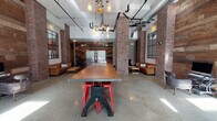 This single-floor communal space masterfully blends industrial and contemporary design elements to create a warm yet sophisticated environment. The interior is anchored by prominent exposed brick columns that rise toward high ceilings, where Edison bulb pendant fixtures and visible ductwork reinforce the space's industrial character.

Polished concrete floors stretch throughout, reflecting natural light that pours in through large, steel-framed windows along the perimeter. Reclaimed wood plank accent walls provide a rich textural contrast to the brick, adding warmth and depth to the overall aesthetic.

The main gathering area features a large communal wood-top table set on a bold red industrial crank base, serving as a natural centerpiece. Multiple lounge seating areas are arranged throughout the space, each appointed with tufted leather Chesterfield sofas, wingback chairs, and reclaimed wood coffee tables — ideal for both collaboration and relaxation.

Near the rear, stainless steel elevator doors are framed by a dramatic reclaimed wood wall, alongside a coffee and amenity station. Large industrial-style mirrors adorn the walls, enhancing the sense of depth and openness. The entry features glass double doors with a branded welcome mat, completing the polished yet unpretentious atmosphere of this thoughtfully designed communal space.