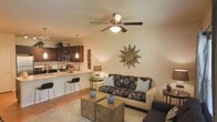 This single-floor residence showcases an open-concept layout that fluidly connects the kitchen and living area, creating a cohesive and functional living environment. The kitchen features rich dark-wood cabinetry, stainless steel appliances, and a light-toned breakfast bar countertop with bar stool seating — an ideal setup for casual dining or entertaining. A pendant light fixture adds a focused, ambient glow above the bar area.

The living room is anchored by a patterned sofa accented with decorative pillows in neutral and blue tones, complemented by rustic wooden coffee tables that add a natural, grounded element to the space. A ceiling fan with an integrated light fixture provides both air circulation and overhead illumination. Warm beige walls and wood-toned flooring contribute to the inviting atmosphere throughout.

A decorative sunburst mirror serves as a notable wall accent, adding visual interest and a touch of artisan character to the living area. A table lamp and side table further enhance the cozy, layered lighting of the room. Overall, the space balances practicality with thoughtful design details.