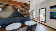 Located at 12400 Toscana Way in Charlotte, NC, this single-floor amenity space showcases a thoughtfully designed interior that blends rustic warmth with contemporary style. The open-plan layout flows seamlessly across wide-plank hardwood floors, connecting several distinct functional areas within a single cohesive environment.

A standout architectural feature is the dramatic slatted wood partition wall, crafted with horizontal timber strips and an inset mountain silhouette upholstered in deep navy leather. This element serves as a natural divider between the lounge and main gathering areas, providing intimate banquette seating alongside marble-top tables beneath elegant pendant lighting.

The main recreation area centers around a full-size billiards table and opens to a generous lounge space with casual seating, creating an inviting atmosphere for social gatherings. Vibrant teal map-inspired wall art and bold artwork panels add artistic energy throughout the space.

A built-in kitchenette features warm wood cabinetry, floating shelves, a matte black sink fixture, and a teal tile backsplash, all illuminated by large black-framed windows that flood the area with natural light and outdoor views. A long communal dining table anchored by cluster pendant lighting accommodates group dining or meetings with ease.

The coffered ceiling detail in the lounge area, combined with recessed lighting and curated décor, gives this amenity space a refined, welcoming character ideal for community use.