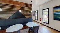 Located at 12400 Toscana Way in Charlotte, NC, this single-floor amenity space showcases a thoughtfully designed interior that blends rustic warmth with contemporary style. The open-plan layout flows seamlessly across wide-plank hardwood floors, connecting several distinct functional areas within a single cohesive environment.

A standout architectural feature is the dramatic slatted wood partition wall, crafted with horizontal timber strips and an inset mountain silhouette upholstered in deep navy leather. This element serves as a natural divider between the lounge and main gathering areas, providing intimate banquette seating alongside marble-top tables beneath elegant pendant lighting.

The main recreation area centers around a full-size billiards table and opens to a generous lounge space with casual seating, creating an inviting atmosphere for social gatherings. Vibrant teal map-inspired wall art and bold artwork panels add artistic energy throughout the space.

A built-in kitchenette features warm wood cabinetry, floating shelves, a matte black sink fixture, and a teal tile backsplash, all illuminated by large black-framed windows that flood the area with natural light and outdoor views. A long communal dining table anchored by cluster pendant lighting accommodates group dining or meetings with ease.

The coffered ceiling detail in the lounge area, combined with recessed lighting and curated décor, gives this amenity space a refined, welcoming character ideal for community use.