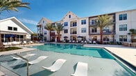 This outdoor amenity area is defined by its resort-inspired design, centered around a generously sized swimming pool with crystal-clear turquoise water. The pool deck is finished in light-toned pavers and lined with modern white in-water lounge chairs, allowing residents to relax directly in the shallow water. Additional poolside chaise lounges and navy blue market umbrellas are arranged throughout the surrounding deck, creating multiple comfortable seating areas ideal for sun and shade.

The pool area is framed by tall, mature palm trees that reinforce the tropical, leisure-focused atmosphere. A separate spa or wading feature is visible adjacent to the main pool, adding further functionality to the outdoor space.

Flanking the pool is a multi-story building showcasing a contemporary coastal farmhouse aesthetic, featuring white board-and-batten siding, dark-framed windows, exposed wood beam accents, and private balconies on upper levels. A covered patio area with exposed beam ceilings, square white columns, and French doors connects interior and exterior spaces seamlessly. Landscaping with native grasses and shrubs completes the grounds, while the overall layout — captured in aerial imaging — reveals a well-organized, U-shaped courtyard configuration designed to maximize communal outdoor living.