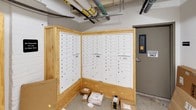 This mail room serves as the central hub for mail and package distribution within the building. The space is defined by its multiple banks of white, individually numbered mailboxes mounted within light natural wood enclosures arranged in an angled configuration, maximizing accessibility and capacity. The mailboxes are organized numerically, allowing residents or occupants to easily locate their assigned units. Each bank accommodates a substantial number of individual compartments, with parcel lockers integrated into the units for oversized deliveries.

The room features polished concrete flooring, white painted walls, and an exposed ceiling with visible utility pipes and conduits running overhead, giving the space a utilitarian, functional character. A secure entry door with an electronic access panel controls entry, ensuring that only authorized personnel can access the area. Signage throughout reinforces this access restriction. Additional open floor space along the walls accommodates larger packages and deliveries that cannot fit within standard mailbox compartments. The overall layout is straightforward and practical, designed to efficiently handle the mail and package needs of the building's occupants.