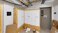 This mail room serves as the central hub for mail and package distribution within the building. The space is defined by its multiple banks of white, individually numbered mailboxes mounted within light natural wood enclosures arranged in an angled configuration, maximizing accessibility and capacity. The mailboxes are organized numerically, allowing residents or occupants to easily locate their assigned units. Each bank accommodates a substantial number of individual compartments, with parcel lockers integrated into the units for oversized deliveries.

The room features polished concrete flooring, white painted walls, and an exposed ceiling with visible utility pipes and conduits running overhead, giving the space a utilitarian, functional character. A secure entry door with an electronic access panel controls entry, ensuring that only authorized personnel can access the area. Signage throughout reinforces this access restriction. Additional open floor space along the walls accommodates larger packages and deliveries that cannot fit within standard mailbox compartments. The overall layout is straightforward and practical, designed to efficiently handle the mail and package needs of the building's occupants.