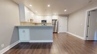 Located at 15267 Hesperian Blvd in San Leandro, California, this single-floor residence offers a thoughtfully updated interior with a clean, contemporary aesthetic throughout. The open-concept kitchen serves as a centerpiece of the space, featuring crisp white shaker cabinetry with brushed nickel hardware, light stone countertops, a peninsula island, and a full suite of stainless steel appliances including a range, over-the-range microwave, dishwasher, and refrigerator. Recessed lighting illuminates the kitchen and living areas, which are finished with rich, dark hardwood-style flooring that extends throughout the main living spaces. The living area benefits from a large sliding glass door dressed with vertical blinds, allowing natural light to filter in and providing access to an outdoor area with views of surrounding greenery. The bedrooms are finished with plush neutral carpeting and include windows with horizontal blinds offering garden views and baseboard heating. The bathroom is well-equipped with a white shaker-style vanity, stone countertop, mirrored medicine cabinet, and a window for natural ventilation. Neutral gray tones on the walls, white trim, and consistent finishes throughout give the entire space a cohesive and move-in-ready appearance.