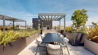 This rooftop terrace is a thoughtfully designed outdoor space that combines contemporary architectural elements with functional amenities and abundant greenery. The terrace is accessed via a walkway flanked by sleek steel horizontal railings and elevated planters filled with ornamental grasses and trees, setting an immediate tone of modern sophistication.

The space is organized into several distinct zones. A dedicated dining area features a large rectangular table surrounded by cushioned rope-woven chairs, positioned beneath a modern louvered pergola. Adjacent to this is a fully equipped outdoor kitchen with stainless steel grills and cabinetry, anchored beneath its own pergola structure. A separate lounging zone offers curved, modular upholstered seating arranged beneath an additional louvered shade structure, ideal for relaxed gathering.

Throughout the terrace, generously sized metal planters house a diverse array of drought-tolerant plants, ornamental grasses, shrubs, and small trees, creating a lush, garden-like atmosphere. Large-format light stone pavers extend across the entire deck, providing a clean, cohesive foundation. The open perimeter, defined by horizontal cable and steel railings, allows unobstructed panoramic views of the surrounding urban landscape beneath open skies.