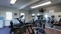 This fitness room is designed to support a well-rounded cardio workout routine in a compact, efficiently arranged space. The room houses a selection of cardio equipment, including elliptical trainers, a stationary bike, and a treadmill, all positioned to make the most of the available floor area. A large wall mirror spans much of one side of the room, visually expanding the space and allowing users to monitor their form during exercise. A wall-mounted television is positioned at a convenient viewing height, providing entertainment during workouts. The room is finished with a durable, dark-toned carpet suited for a fitness environment, while the light gray walls contribute to a clean, neutral aesthetic. Overhead fluorescent lighting ensures the space is well-lit throughout. Windows along the walls allow natural light to filter in, adding to the openness of the room. Framed artwork on the walls adds a modest decorative touch. The overall layout is practical and straightforward, making this fitness room a convenient and functional amenity for regular use.