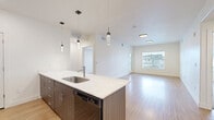 Located at 450 S State St in Clearfield, Utah, this well-appointed 2-bedroom, 2-bathroom unit offers a thoughtfully designed single-floor layout. The open-concept kitchen and living area serves as the heart of the home, showcasing a sleek kitchen island with a white quartz countertop and undermount sink, complemented by stylish pendant lighting overhead. Dark wood-toned lower cabinetry contrasts elegantly with light gray upper cabinets, and black stainless steel appliances add a modern edge. Warm wood-style flooring runs throughout the main living areas, creating a cohesive and inviting atmosphere. The living room benefits from natural light streaming through large windows with outdoor views. Both bedrooms feature plush carpeting and generous windows, with the primary bedroom offering a tray ceiling detail. A dedicated closet provides ample hanging and storage space. The bathrooms are clean and functional, featuring wood-toned vanities with white countertops and a tub-shower combination finished with brushed nickel fixtures. A dedicated laundry area with in-unit washer hookups and shelving adds everyday convenience. The hallway connects all spaces efficiently, contributing to the unit's practical and modern feel throughout.