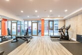 Located at 1424 Highland Rd W in Kitchener, Ontario, this fitness facility offers a well-equipped workout space within a single open-concept floor. The room's design combines functionality with a modern aesthetic, highlighted by bold orange accent walls that contrast with a concrete-effect feature wall and crisp white ceilings fitted with recessed pot lighting.

The flooring transitions between light wood-style vinyl in the central zone and durable rubber matting in the equipment areas, providing both visual appeal and practical protection. A floor-to-ceiling glass window and door frontage faces the street, flooding the space with natural light and offering clear views to the exterior.

The facility is outfitted with a variety of equipment to support diverse training needs. Cardio options include a treadmill and a stationary bike, while strength training is supported by a multi-function cable/pulley machine, an adjustable weight bench, and a well-stocked dumbbell rack with a full range of free weights. A full-length wall mirror enhances the sense of openness and allows users to monitor their form during workouts.

A dedicated entry corridor connects to the main workout area, with a wall-mounted split HVAC unit ensuring year-round climate comfort. The overall layout is efficient and thoughtfully organized, making effective use of the available floor area to deliver a comprehensive amenity fitness space.