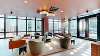 Located at 215 Grove St in Jersey City, NJ, this striking single-floor space offers a generous open-plan layout that seamlessly blends a sophisticated lounge, a fully equipped kitchen, and remarkable outdoor terrace access. The living area is anchored by dramatic floor-to-ceiling glass walls that flood the interior with natural light and frame panoramic views of the Jersey City skyline. A warm terracotta-toned ceiling adds a bold architectural accent, complemented by sculptural pendant lighting and a statement ceiling fixture. The lounge is furnished with a curated mix of leather sofas and rounded upholstered chairs arranged around low-profile coffee tables, creating an inviting yet design-conscious atmosphere. The integrated kitchen features a substantial wood-finished island, blush-toned cabinetry, and professional-grade appliances, making it as functional as it is visually striking. The outdoor terraces extend the living space considerably, offering tiled open-air areas with lounge chairs, a large communal dining table with vibrant red seating, and glass railings that maximize unobstructed views of the surrounding cityscape and beyond. A partially covered terrace section with a distinctive circular skylight overhead adds an architectural focal point, enhancing the sense of refinement throughout this exceptional urban space.