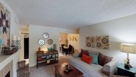 This single-floor residence offers a comfortable and inviting layout designed for everyday living. The living area serves as the heart of the home, featuring plush wall-to-wall carpeting that adds warmth underfoot. A neutral color palette on the walls provides a versatile backdrop, complemented by decorative wall art and eclectic accents that lend the space a personalized, lived-in charm. A cushioned sofa with colorful throw pillows anchors the seating area, paired with a rich dark wood coffee table that grounds the room. An open passage connects the living area to a separate dining space, which features a dark wood dining table and chairs set against warm-toned walls, creating a distinct yet cohesive entertaining zone. A wooden console table along the living room wall provides additional display and storage space, showcasing decorative items that add character. The textured popcorn ceiling adds a classic touch throughout. The open sightlines between rooms promote a sense of flow and connectivity, making the space feel both functional and welcoming for everyday living and casual entertaining.