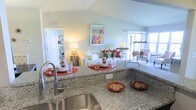 This single-floor residence offers a thoughtfully designed open-concept layout that seamlessly connects the kitchen, dining, and living areas. The kitchen is a standout feature, appointed with rich dark cabinetry, stainless steel appliances, and a generous granite countertop island with bar seating — ideal for casual dining and entertaining. A stainless steel undermount sink and sleek fixtures complete the space. The kitchen flows directly into the dining area, which is anchored by a stylish table set beneath a pendant light and a striking round mirror that adds depth and elegance to the wall. Vaulted ceilings carry through the main living area, enhancing the sense of openness and airiness. The living room is bright and welcoming, with soft neutral tones and warm lighting creating an inviting atmosphere. A sunroom or enclosed sitting area at one end of the residence is wrapped in large multi-pane windows that flood the space with natural light and offer open outdoor views, making it a perfect retreat. Throughout the home, neutral wall tones, plush carpeting, and tile flooring create a cohesive and comfortable aesthetic. The overall ambiance is warm, polished, and move-in ready.