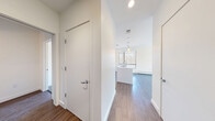 Located at 110 Winooski Falls Way in Winooski, Vermont, this thoughtfully designed 2-bedroom, 2-bathroom residence offers a clean, contemporary aesthetic throughout its single-floor layout. The open-concept kitchen and living area serves as the heart of the home, featuring sleek, textured cabinetry in a soft gray-taupe finish, stainless steel appliances including a gas range and built-in microwave, and a crisp white island countertop with an undermount sink. Pendant lighting and large windows fill the kitchen with natural light and offer views of the surrounding treeline. Hardwood-style flooring runs through the main living areas, complementing the bright white walls and modern trim. The spacious primary bedroom provides generous proportions, plush carpeting, and a large window framing wooded scenery. The second bedroom, also carpeted, offers a quiet, versatile space. One bathroom features a floating vanity with a vessel sink, gray wood-tone cabinetry, and a large mirror with wall sconces, while the second includes a full tub-shower combination with earthy tile surround. A wall-mounted HVAC unit and a private outdoor access door enhance everyday comfort and convenience. The hallway connects all spaces efficiently, completing this well-appointed modern residence.