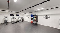 Located at 1701 Federal St in Philadelphia's Graduate Hospital neighborhood, this fitness center offers a well-organized and fully equipped workout space designed to support a wide range of training needs. The interior features crisp white walls adorned with motivational typography decals — phrases such as "Stay Positive," "Work Hard," and "Don't Stop Until You're Done" — creating an energizing atmosphere throughout the space. Durable dark rubber flooring covers the entire area, providing a safe and functional surface for all types of exercise. The ceiling features exposed black piping alongside bright linear LED lighting, lending the space an industrial-modern aesthetic. Cardio equipment includes multiple treadmills and an elliptical machine, each equipped with individual monitors. Strength training is well-supported with a multi-function cable machine, an adjustable workout bench, a full rack of dumbbells, and an assortment of kettlebells and medicine balls on a dedicated storage rack. A large wall-mounted mirror expands the sense of space and aids in form correction during workouts. A mounted flat-screen TV, a wall-mounted split air conditioning unit, and access to two separate doors — likely leading to restroom or utility areas — round out the practical amenities of this clean and functional fitness facility.