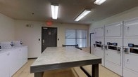 This single-floor laundry facility is designed with practicality and functionality in mind. The room is illuminated by overhead fluorescent lighting that ensures consistent brightness throughout the space. A prominent feature is the large, flat folding table positioned centrally, providing ample surface area for sorting and folding laundry. Along the right wall, a row of stacked washer and dryer units offers convenient, efficient use of space, each equipped with clearly visible control panels. The room features neutral-toned walls and a textured ceiling, contributing to a clean, utilitarian aesthetic. A window fitted with horizontal blinds allows for natural light while maintaining privacy. The space includes a clearly marked emergency exit door, reinforcing safety compliance. The overall layout is open and accessible, making it easy to navigate while managing laundry tasks. The no-smoking signage posted near the entrance further emphasizes the facility's commitment to a safe and comfortable environment for all users.