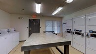 This single-floor laundry facility is designed with practicality and functionality in mind. The room is illuminated by overhead fluorescent lighting that ensures consistent brightness throughout the space. A prominent feature is the large, flat folding table positioned centrally, providing ample surface area for sorting and folding laundry. Along the right wall, a row of stacked washer and dryer units offers convenient, efficient use of space, each equipped with clearly visible control panels. The room features neutral-toned walls and a textured ceiling, contributing to a clean, utilitarian aesthetic. A window fitted with horizontal blinds allows for natural light while maintaining privacy. The space includes a clearly marked emergency exit door, reinforcing safety compliance. The overall layout is open and accessible, making it easy to navigate while managing laundry tasks. The no-smoking signage posted near the entrance further emphasizes the facility's commitment to a safe and comfortable environment for all users.
