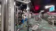 This single-floor fitness facility in Berkeley, California, delivers a visually dynamic and fully equipped workout environment. The interior design makes an immediate impression through a combination of dark charcoal walls, a matte black exposed ceiling with industrial ductwork, and a vivid teal-and-grey patterned carpet that runs throughout the space. Tropical palm-print wallcoverings line the primary accent wall, complemented by bold black-and-white artistic panels, creating an energetic aesthetic reinforced by a bright pink neon sign reading "Work Hard, Play Hard."

Lighting is a key design element, with suspended linear fixtures providing functional illumination while pink-hued accent lighting at the ceiling adds atmosphere. Full-length mirrors along one wall visually expand the space and support functional training.

The facility is comprehensively equipped for diverse workout goals. A cable machine and adjustable bench anchor the strength-training zone, accompanied by a full rack system holding kettlebells, medicine balls, stability balls, and battle ropes. A dumbbell rack and additional free weights are accessible along the mirrored wall. The cardio area includes stair climbers and other aerobic machines. Rolled SmartMats and foam rollers are available for stretching and floor work. A monitor mounted near the ceiling adds a media component to the training environment.