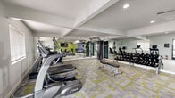 This fitness center at 2800 S Mission Rd in Tucson, Arizona, offers a well-appointed workout space designed to support a comprehensive exercise routine. The single-floor layout is thoughtfully organized to maximize usability while maintaining an open, spacious feel. Flooring throughout features a modern geometric carpet tile in neutral gray and yellow-green tones, adding a vibrant yet functional aesthetic to the space. Walls are finished in crisp white, with an accent wall in a fresh sage green that adds energy and visual interest. Large mirrors line key sections of the walls, enhancing the sense of depth and allowing users to monitor their form during workouts. The cardio zone is equipped with TRUE brand elliptical trainers and treadmills, as well as a rowing machine and a stationary spin bike with its own interactive display. A multi-functional cable machine anchors the strength training area, complemented by a full dumbbell rack and an adjustable weight bench. Recessed lighting and a ceiling fan ensure the space remains well-lit and comfortable throughout use. A wall-mounted flat-screen TV adds an entertainment option during workouts. Natural light filters in through windows, and the facility includes a dedicated entryway. Informational and safety signage is neatly posted throughout the space.