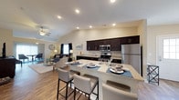 This single-story space is designed with an open-concept layout that connects the kitchen, dining, and living areas into one cohesive environment. The kitchen is a standout feature, equipped with stainless steel appliances including a refrigerator, range, and built-in microwave. Rich, dark-toned cabinetry extends along the full length of the kitchen wall, providing ample storage, while light granite countertops offer a striking contrast. A large kitchen island with matching granite serves as both a functional workspace and a casual dining area, accommodating multiple bar-height seating positions with modern upholstered stools.

The vaulted ceiling enhances the sense of openness throughout the space, with recessed lighting providing consistent illumination. Warm-toned hardwood-style flooring runs throughout, adding cohesion between zones. The adjacent living area features a ceiling fan, neutral walls, and large windows that allow natural light to fill the room. The overall aesthetic balances contemporary finishes with a comfortable, functional layout suited for everyday living and entertaining.