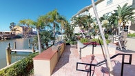 Located at 14439 Compass St in Corpus Christi, Texas, this outdoor amenity space offers a vibrant, resort-style environment designed for relaxation and recreation. The grounds are richly landscaped with tall palm trees, tropical plantings, and colorful seasonal flowers, creating a lush coastal atmosphere throughout.

A stamped concrete patio serves as the central gathering area, featuring multiple seating zones with comfortable chairs and tables arranged beneath shaded pergola structures. The outdoor kitchen is a standout feature, equipped with a full-sized stainless steel grill and ample counter space, making it ideal for outdoor entertaining and cookouts.

The resort-style swimming pool is a focal point of the space, surrounded by lounge chairs and bordered by manicured greenery and swaying palms under wide-open Texas skies. Adjacent to the pool, a covered area provides shaded retreat space with additional seating and recreational amenities, including an oversized outdoor Jenga game.

A waterfront patio area offers direct views of the marina and surrounding water, with a dock visible in the background, underscoring the property's prime coastal location. Wrought iron fencing frames the perimeter of the outdoor space, providing security while preserving the open, airy feel. The entire area benefits from abundant natural sunlight and refreshing Gulf Coast breezes, delivering a relaxed, vacation-inspired ambiance year-round.