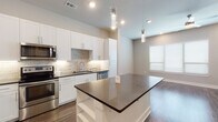 This single-floor residence showcases a thoughtfully designed open-concept layout, where the kitchen flows seamlessly into the living area. The kitchen is a standout feature, equipped with crisp white shaker-style cabinetry extending to the ceiling for generous storage, dark countertops, and a full suite of stainless steel appliances including a refrigerator, range, microwave, and dishwasher. A textured stone tile backsplash adds visual depth, while under-cabinet lighting enhances the workspace. A central island with a matching dark countertop provides additional prep space and serves as a natural gathering point. Cylindrical pendant lights hang above the island, adding a contemporary touch. Rich wood-look flooring runs throughout the main living areas, complementing the neutral color palette. The living area benefits from natural light through blinded windows. The bedrooms feature plush carpeting and large windows fitted with horizontal blinds. The bathroom is finished with a tiled tub-and-shower combination featuring horizontal stripe detailing, a white vanity with dark countertop, a framed mirror, and coordinating tile flooring, creating a clean and cohesive aesthetic throughout.