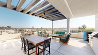 Located at 4212 Genesee Ave in San Diego, California, this rooftop deck — named the Grace Hawkes Rooftop Deck — offers an expansive open-air retreat designed for both relaxation and social gatherings. The deck is defined by a striking pergola structure with bold, evenly spaced overhead beams that provide partial shade while framing sweeping panoramic views of the San Diego skyline, surrounding neighborhoods, and distant mountain ranges.

The space is thoughtfully divided into distinct functional zones. A lounge area features wood-framed seating with teal cushions arranged around a circular table, creating a comfortable and inviting setting for casual conversation. Nearby, a generous dining area centers on a large wooden table surrounded by slatted chairs, ideal for group meals and events. Additional bistro-style seating with compact round tables is positioned along the perimeter, offering more intimate spots to enjoy the open-air setting.

An outdoor kitchenette area with counter space and a wall-mounted television adds convenience and entertainment value to the space. The crisp white exterior walls contrast cleanly with the warm wood furnishings and the vibrant teal upholstery, lending the deck a modern yet welcoming aesthetic. Bathed in abundant Southern California sunlight and open to refreshing breezes, this rooftop deck delivers a premium outdoor experience year-round.