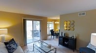 This single-floor residence offers a thoughtfully designed open-concept layout that seamlessly connects the living and dining areas, creating a cohesive and inviting atmosphere. The living room is anchored by plush carpeting and features a glass-top coffee table set atop a patterned area rug, providing a clean and modern aesthetic. Warm golden-yellow walls lend a welcoming energy throughout the space, complemented by neutral ceilings that keep the overall feel light and airy. A large sliding glass door floods the living area with natural light and opens directly onto a private balcony, where outdoor seating offers a pleasant retreat overlooking mature trees. The dining area is defined by an elegant chandelier that adds a refined touch, and is furnished with a dark-toned dining set suited for intimate gatherings. A window beside the dining table draws in additional natural light. Along the living room wall, a dark media console provides generous storage and display space, accented by a stylish circular mirror arrangement that adds depth and visual interest. The combination of warm tones, natural light, and functional furnishing areas makes this a comfortable and well-appointed living environment.
