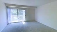 This single-floor residence offers a clean, functional layout designed for comfortable everyday living. The living area is finished with wall-to-wall carpeting that adds warmth underfoot, while a large sliding glass door floods the room with natural light and opens onto a private balcony overlooking mature trees — creating a serene, nature-connected atmosphere. The neutral, light-colored walls throughout contribute to a bright and airy feel.

The kitchen is thoughtfully laid out with white cabinetry providing generous storage space above and below the countertops. White appliances, including a freestanding electric range and a full-size refrigerator, are neatly integrated into the workspace. Tile flooring in the kitchen provides a practical, easy-to-clean surface. A half-wall partition separates the kitchen from the adjacent hallway, lending a sense of structure while keeping the space open.

A carpeted hallway connects the main living areas to the rest of the unit, with flush-mounted ceiling fixtures providing consistent lighting throughout. Doors along the hallway suggest well-defined, private spaces within the home. Overall, the unit presents a tidy, move-in ready environment with a straightforward, practical design aesthetic suited for a variety of lifestyles.
