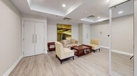 This thoughtfully designed single-floor amenity space offers a generous and versatile layout ideal for both social gatherings and quiet meetings. The space is defined by elegant tray ceilings with recessed lighting throughout, creating a polished and refined atmosphere. Light wood-style flooring flows seamlessly across the entire floor, unifying the distinct functional zones.

A central wet bar with a dark stone countertop and warm wood cabinetry serves as a focal point, complemented by bar seating and convenient access to the surrounding areas. Adjacent to the bar, a comfortable lounge area features neutral-toned sofas and accent chairs arranged around a rich mahogany coffee table, with a bold abstract painting anchoring the wall. Double white doors provide access to additional rooms or storage.

Multiple dining areas are distributed throughout the space, each furnished with rich wood tables and upholstered chairs. These dining nooks benefit greatly from large floor-to-ceiling windows that frame expansive urban skyline views, flooding the space with abundant natural light. Wall-mounted flat-screen televisions add a modern, functional touch. The overall ambiance is clean, contemporary, and inviting — well-suited for community use in a residential or hospitality setting.