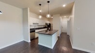 This well-appointed single-floor residence offers a thoughtfully designed open-concept layout that seamlessly connects the kitchen and living areas. The kitchen is a standout feature, showcasing crisp white shaker-style cabinetry paired with striking dark granite countertops that extend across both the perimeter surfaces and a central island. Pendant globe lights hang above the island, adding a contemporary touch, while stainless steel appliances — including a refrigerator, microwave, and range — complete the functional and stylish space. Rich, dark hardwood flooring flows throughout the main living areas, creating a warm and cohesive aesthetic. The living room is bright and open, benefiting from natural light through a glass-paneled door and a ceiling fan overhead, lending the space a comfortable, airy feel. The bedrooms are carpeted for added comfort, each featuring ceiling fans and windows that allow natural light to filter in. The bathroom is finished with classic white subway tile surrounding a tub-shower combination and chrome fixtures. A hallway connects the rooms efficiently, and the overall neutral color palette of warm whites and beiges creates a clean, versatile backdrop throughout the entire space.