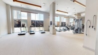 Located at 850 Pacific St in Stamford, Connecticut, this generously sized fitness center spans a single floor and offers a comprehensive workout environment designed to accommodate a wide range of exercise needs. The main gym area is bright and open, featuring soaring ceilings with warm wood-accented linear pendant lighting that adds a modern yet inviting aesthetic. Large windows along the perimeter flood the space with natural light and provide views of the surrounding neighborhood, creating an energizing atmosphere. The main floor is divided into distinct functional zones: a cardio section lined with treadmills and elliptical machines, a strength training area equipped with cable machines, multi-functional weight systems, and an extensive selection of free weights including dumbbells and barbells with adjustable benches. A dedicated free weights zone features bold gray and yellow accent walls with rubber flooring for durability and safety. A separate studio room offers a quieter, more versatile setting with wall-to-wall mirrors, ballet barres, TRX suspension equipment, yoga mats, and a large display screen — ideal for yoga, barre, or group fitness classes. The entry to the facility features double glass doors with clerestory windows above, contributing to the space's airy, open feel throughout.