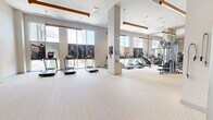 Located at 850 Pacific St in Stamford, Connecticut, this generously sized fitness center spans a single floor and offers a comprehensive workout environment designed to accommodate a wide range of exercise needs. The main gym area is bright and open, featuring soaring ceilings with warm wood-accented linear pendant lighting that adds a modern yet inviting aesthetic. Large windows along the perimeter flood the space with natural light and provide views of the surrounding neighborhood, creating an energizing atmosphere. The main floor is divided into distinct functional zones: a cardio section lined with treadmills and elliptical machines, a strength training area equipped with cable machines, multi-functional weight systems, and an extensive selection of free weights including dumbbells and barbells with adjustable benches. A dedicated free weights zone features bold gray and yellow accent walls with rubber flooring for durability and safety. A separate studio room offers a quieter, more versatile setting with wall-to-wall mirrors, ballet barres, TRX suspension equipment, yoga mats, and a large display screen — ideal for yoga, barre, or group fitness classes. The entry to the facility features double glass doors with clerestory windows above, contributing to the space's airy, open feel throughout.