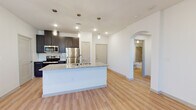 This single-floor residence showcases a clean, contemporary design with an open-concept layout that seamlessly connects the kitchen and living areas. Light wood-style flooring extends throughout the main living spaces, creating a bright and cohesive atmosphere, while recessed lighting and pendant fixtures provide well-distributed illumination.

The kitchen is a functional centerpiece, anchored by a spacious island with granite countertops, an undermount double sink, and dark espresso cabinetry with brushed nickel hardware. Stainless steel appliances — including a refrigerator, range, and microwave — add a polished, modern touch, complemented by a tile backsplash. Pendant lights hang above the island, reinforcing the kitchen's role as a gathering space.

The open living area is generously proportioned and benefits from natural light, with white walls enhancing the sense of airiness. An elegant arched doorway leads to the bathroom, which features a granite-topped vanity with dark cabinetry, a large framed mirror, and a bathtub. A separate carpeted bedroom offers a quieter, softer environment. Additional interior doors provide access to closets and utility spaces, rounding out this well-appointed residence.