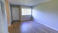 This single-floor space offers a straightforward and functional layout designed to maximize usability. The main living area is finished with durable wood-look plank flooring in a warm medium-tone hue, providing a clean and cohesive aesthetic throughout. Neutral gray walls create a versatile backdrop that complements a wide range of furnishing styles and décor choices.

A large picture window serves as a prominent feature of the main room, drawing in natural light and framing views of mature trees and the surrounding outdoor environment. The window enhances the sense of openness within the space, making it feel bright and airy. White baseboards provide a crisp contrast against the gray walls and wood-tone flooring, contributing to a polished, well-maintained appearance.

The entry door is finished in white and includes a light switch panel nearby, offering a practical and clearly defined entryway. The ceiling features a textured popcorn finish, which adds subtle dimension overhead. Electrical outlets are conveniently positioned along the walls. Overall, the space presents a clean, neutral, and move-in ready environment suitable for a variety of uses and furnishing configurations.