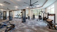 Located at 30000 Summer Crop Lane in Apex, NC, this expansive fitness center offers a comprehensive and well-appointed exercise facility across a single open floor. The space is designed with a modern aesthetic, featuring tray ceilings accented with warm crown molding, oversized industrial-style ceiling fans for air circulation, and recessed lighting throughout. Patterned, multi-tone carpet tiles cover the floors, adding both texture and durability suited for a high-traffic fitness environment. Large floor-to-ceiling windows line the perimeter walls, flooding the space with abundant natural light and providing pleasant views of the surrounding wooded landscape. The cardio zone is well-equipped with multiple treadmills, elliptical machines, and spin bikes, all positioned to take advantage of the outdoor views. A dedicated strength training area houses a variety of cable machines, multi-functional racks, weight benches, and plate-loaded equipment. A functional training zone provides plyometric boxes, medicine balls, stability balls, and an extensive wall-mounted storage rack with kettlebells, resistance bands, and free weights. The layout is thoughtfully organized to allow ample space between equipment zones, promoting safe and efficient movement throughout the facility.