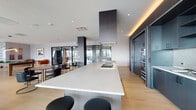 This thoughtfully designed single-floor space combines modern aesthetics with versatile functionality across a generously proportioned open layout. Light hardwood flooring flows seamlessly throughout, unifying each distinct zone while recessed lighting and warm cove accents enhance the refined atmosphere.

The centerpiece of the kitchen is a striking oversized island with a light stone countertop, built-in cooktop, and a bold geometric range hood suspended above. Dark, floor-to-ceiling cabinetry lines the perimeter wall, housing integrated appliances, open shelving, a tile backsplash, and a dedicated sink station — all finished with sleek hardware for a polished, contemporary look.

The lounge area features plush teal velvet sectional sofas anchored by a textured, metallic accent wall with an integrated media display. Nearby seating vignettes with cognac leather chairs and curated wall art provide intimate gathering spots, while sculptural decorative elements add artistic character throughout.

The game room and social zone open dramatically to the outdoors through full-height glass walls and doors, flooding the space with natural light and offering sweeping panoramic city views. Pool tables, casual seating clusters, and flexible dining arrangements make this area ideal for both relaxed recreation and social events.

Glass-paneled interior partitions and dark stone column accents define transition areas, lending an architectural sophistication that ties the entire space together.