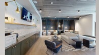 Located at 110 Addison Dr in Athens, GA, this thoughtfully designed single-floor amenity space blends modern aesthetics with functional versatility. The main lounge area sets a welcoming tone with light hardwood flooring, a distinctive slatted ceiling with track lighting, and a wall-mounted TV flanked by warm brass sconce lighting. A sleek built-in credenza with dark green cabinetry and brass hardware runs along one wall beneath a textured tile backsplash, providing both storage and display space. Comfortable seating arrangements, including a neutral sofa, a bold navy accent chair, and a patterned area rug, create an inviting social hub centered around a substantial coffee table. Semi-private dining booths line the back wall, upholstered in cream boucle fabric set against deep charcoal paneling, offering more intimate settings for small gatherings or focused work. A bright, windowed conference room features floor-to-ceiling steel-framed windows with sheer drapery, flooding the space with natural light and outdoor views. A statement gold pendant light anchors a long wood conference table surrounded by leather chairs. An additional flex lounge area accessed through large sliding barn doors offers casual seating and its own generous window wall, making it ideal for informal meetings or relaxation. Throughout, the design balances sophistication with comfort.