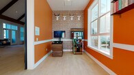 Located at 2200 Silver Crescent Dr in Charlotte, NC, this single-floor community amenity space blends energetic design with comfortable social areas. The interior is defined by bold, warm orange walls accented with crisp white wainscoting and light hardwood-style flooring that runs throughout, creating a cohesive and lively atmosphere.

The space is divided into two distinct zones. The game area features an eye-catching patterned accent wall adorned with decorative deer head mounts, housing an arcade cabinet and a tabletop gaming console. This area benefits from a large window that allows ample natural light to pour in, offering views of the surrounding outdoor environment.

Adjacent to the game area, the lounge zone is bathed in abundant natural light streaming through oversized windows that span much of the wall. Built-in window bench seating with decorative throw pillows provides a relaxed setting, complemented by plush green velvet accent chairs arranged around a sculptural pedestal table. A cowhide-style area rug grounds the seating arrangement, adding texture and contrast. Wall-mounted shelving and bold domino-inspired artwork contribute to the playful, curated aesthetic.

The open layout connects seamlessly to a larger gathering area visible through the space, which features soaring vaulted ceilings with dark exposed beams, reinforcing the expansive and welcoming character of this community hub.
