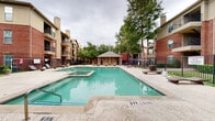 Located at 401 N Old Orchard Ln in Lewisville, Texas, this outdoor pool area serves as a central amenity space within a well-maintained residential community. The centerpiece of the space is a generously sized swimming pool with clear, turquoise water, accompanied by an adjacent spa for relaxation. The pool deck is finished with a smooth concrete surround, providing ample space for lounge chairs and seating arranged around the perimeter for sunbathing and leisure. A charming covered cabana with a pitched roof anchors one end of the courtyard, offering a shaded retreat for residents. The space is enclosed by a black wrought-iron fence, providing both security and an open, airy feel. Mature trees are dispersed throughout the courtyard, offering natural shade and a lush, green backdrop that enhances the overall ambiance. Decorative lamp posts and potted plantings add to the well-kept aesthetic. The pool area is surrounded on multiple sides by multi-story brick residential buildings featuring private balconies, which overlook the courtyard and contribute to the sense of an intimate, community-centered environment. The combination of resort-style amenities, natural greenery, and classic brick architecture creates an inviting and functional outdoor living space.