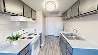 This single-floor space is centered around a thoughtfully designed galley-style kitchen that maximizes functionality within a streamlined layout. The kitchen features slate-blue lower and upper cabinetry with brushed nickel hardware, offering ample storage on both sides of the corridor. Light quartz-style countertops provide generous prep space and contrast neatly with the cabinetry. On one side, a white electric range with coil burners is paired with an over-range ventilation hood and a countertop microwave, making meal preparation convenient and efficient. The opposite side hosts a stainless steel double-basin sink with a modern faucet, flanked by additional cabinetry. A subtle tile backsplash adds texture near the cooking area. Underfoot, wood-look laminate flooring in a warm gray tone runs the length of the kitchen, lending a clean, contemporary feel. A flush-mount ceiling light fixture brightens the space, complemented by a ceiling vent for climate control. At the far end of the kitchen, a set of white bi-fold doors provides access to a closet or utility area, contributing to the overall storage capacity of the space. The light, neutral wall tones keep the interior feeling open and airy.