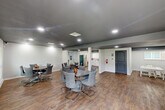 This well-appointed single-floor space offers a thoughtfully designed layout that accommodates a variety of activities and group gatherings. The main area is anchored by multiple round dining tables with modern gray upholstered chairs, providing ample seating for meetings, meals, or social events. Warm wood-look flooring runs throughout, complementing the clean white walls and dark painted ceilings fitted with recessed lighting that creates a bright, welcoming ambiance.

A dedicated activity zone features white tables and stools, ideal for arts, crafts, or educational programming. A wall-mounted flat-screen television adds a media-ready element to the main gathering area. Structural columns are seamlessly integrated into the open floor plan, maintaining a sense of spaciousness throughout.

The kitchenette area is equipped with white cabinetry, a stainless steel refrigerator, and counter space, supporting catering and refreshment needs. A standout feature is the glass-enclosed fitness room, which houses cardio equipment and is lined with mirrors, offering a transparent yet defined space for exercise.

The layout is completed by multiple gray-toned doors providing access to restrooms and utility areas. Natural light filters in through windows, complementing the recessed lighting. Overall, this space delivers a clean, modern, and functional environment suited for community use, events, and wellness activities.