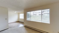 This single-floor space offers a clean, functional layout with a bright and open living area. Large horizontal windows, fitted with horizontal blinds, line one wall and allow generous natural light to fill the room while framing views of surrounding greenery and neighboring buildings. An additional window on the adjacent wall further enhances the sense of airiness. The flooring is covered in neutral-toned carpet, complementing the light beige walls and creating a cohesive, understated aesthetic. A textured popcorn ceiling adds a classic touch overhead, while a simple flush-mount light fixture provides ambient illumination. Baseboard heaters run along the perimeter of the room, offering a practical and consistent heating solution. The open layout of the living area provides flexible space for a variety of furniture arrangements. The overall ambiance is calm and understated, with an emphasis on natural light and simplicity, making it a practical and comfortable single-level space.