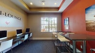 The I-Lounge is a thoughtfully designed, single-floor communal space built to support both productivity and casual interaction. The room is anchored by a long, dark workstation desk lining one wall, equipped with multiple iMac computers and a shared printer — ideal for residents or guests who need reliable access to technology and Wi-Fi. The branded "I-Lounge" signage, complete with a Wi-Fi symbol, is prominently displayed above the workstation area, reinforcing the space's connected, tech-forward identity.

A distinctive tray ceiling with recessed lighting adds architectural depth and a polished finish to the room. The color palette contrasts warm taupe walls with bold terracotta-red accent walls, creating an energetic yet grounded atmosphere. Dark blue-grey carpeting grounds the space while complementing the modern furnishings throughout.

On the lounge side of the room, sleek high-top tables with contemporary white bar stools provide casual seating for collaboration or relaxed use. A vibrant beach-themed wall canvas adds a local, coastal character to the space. Large grid-pane windows allow generous natural light to filter in, offering pleasant views of swaying palm trees outside. A frosted glass interior door connects to an adjacent area, contributing to the space's open, accessible layout.