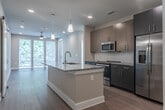 Located in Atlanta's vibrant Glenwood Park neighborhood, this modern single-floor residence offers a thoughtfully designed open-concept layout with contemporary finishes throughout. The kitchen serves as the heart of the home, anchored by a large center island with a granite countertop and undermount sink, illuminated by sleek pendant lighting. Dark cabinetry with stainless steel hardware contrasts beautifully against taupe upper cabinets, while stainless steel appliances — including a refrigerator, range, and built-in microwave — complete the space. Rich hardwood-style flooring flows seamlessly from the kitchen into the adjacent living area, which is bathed in natural light from large floor-to-ceiling windows and sliding glass doors. The bedrooms feature plush carpeting, ceiling fans, and generous windows that frame leafy outdoor views, creating a calm and comfortable retreat. The bathrooms are finished with large-format tile flooring, dark vanities topped with granite countertops, and framed mirrors. One bathroom includes a soaking tub with a tiled surround. A dedicated laundry room with wire shelving and washer/dryer hookups adds practical convenience. A stylish sliding barn door accent adds a touch of modern character to the overall design.