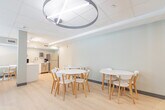 Located at 699 Niagara St in Welland, Ontario, this single-floor communal space is designed to provide a welcoming and functional environment for its users. The layout is efficiently organized to accommodate multiple purposes within a compact footprint, seamlessly blending a lounge area, dining zone, and a fully equipped kitchenette.

The lounge area features rich brown leather sofas arranged around a small white accent table, anchored by a large circular pendant light fixture that adds a modern, polished touch to the drop-tile ceiling. A wall-mounted flat-screen television serves as a central focal point, while natural light filters in through roller-shaded windows, creating a bright and relaxed atmosphere. A dedicated activity table stocked with games and books provides an engaging space for leisure.

The dining area includes a clean white oval table paired with contemporary wood-framed chairs, set against soft sage-green walls and light hardwood-style flooring that flows throughout the space. The kitchenette is well-appointed with stainless steel appliances — including a full-size refrigerator, dishwasher, and microwave — complemented by white cabinetry and a double sink. A glass-paneled door connects to an adjacent office or administrative area, enhancing the functional versatility of this thoughtfully curated space.